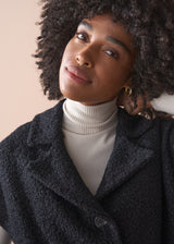 Person wearing a black boucle gilet jacket over a cream poloneck and black trousers against a beige background