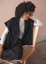 Person wearing a black boucle gilet jacket over a cream poloneck and black trousers against a beige background