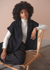 Woman wearing a black boucle gilet jacket over a cream turtleneck, sitting on a wooden chair against a beige background