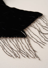 A close-up of a black velvet scarf with silver beaded fringe, laid flat on a light beige textured surface.