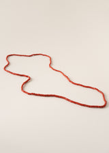 A long beaded necklace made of small red beads, laid out in a loose loop on a plain light beige background.