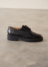 A single black leather oxford shoe with a low block heel and brogue detailing, photographed on a textured gray concrete floor against a neutral beige wall.