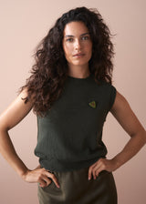 Woman wearing a dark green sleeveless top with a green heart brooch against a beige background