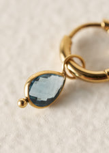 A close-up of a gold hoop earring with a teardrop-shaped light blue gemstone and a small gold bead accent, set against a textured off-white background.
