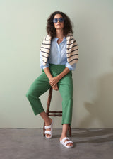 Woman sitting on a wooden stool wearing green trousers, a light blue shirt, and a striped cardigan.