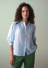 Woman wearing a light blue shirt and green trousers against a plain background