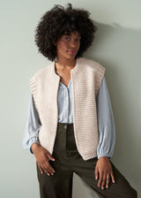 Woman wearing a beige knitted vest over a pale blue shirt and dark green trousers with a neutral background