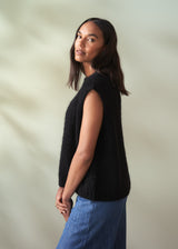 Woman wearing a black sleeveless knit sweater and blue jeans against a neutral background