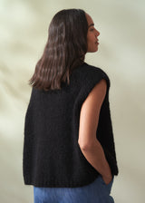Woman wearing a black sleeveless knit sweater and blue jeans against a neutral background