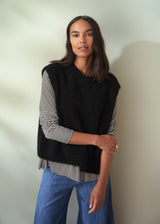 Woman wearing a black sleeveless knit sweater and blue jeans against a neutral background