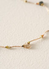 A delicate beaded necklace with a translucent pale pink heart-shaped bead and small green and gold beads on a thin cord, photographed on a textured off-white surface.