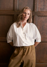A woman with blonde hair stands against a dark brown wooden paneled wall, wearing a white button-down blouse with puff sleeves and brown drawstring trousers.