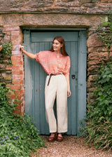 Woman wearing a red checkered shirt with white floaty trousers and brown leather clogs, leant against the doorway of an old stone building
