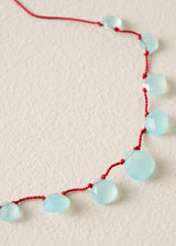 A close-up of a necklace with faceted light blue beads strung on a red cord, laid on a textured off-white surface.