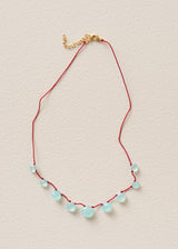 A delicate necklace with a red cord, small light blue faceted beads, and a gold clasp, laid flat on a textured beige background.