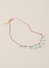 A delicate necklace with a thin red cord, small light blue faceted beads, and a gold clasp, displayed on a textured beige background.