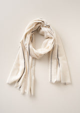 An off-white woven scarf with black dotted trim along the edges, draped in a loose loop against a solid, light beige background.