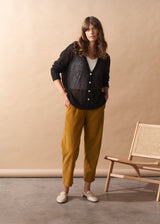 Woman wearing a black cardigan, white shirt, and yellow pants standing against a beige wall with a wooden chair.