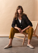 Woman sitting on a chair wearing a black jacket, white shirt, mustard pants, and white loafers.