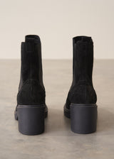 A pair of black suede Chelsea-style ankle boots with chunky block heels, shown from the back against a neutral beige and gray studio background.