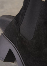 Close-up of a black suede ankle boot with a black elastic side panel and a chunky black block heel, photographed against a neutral gray background.