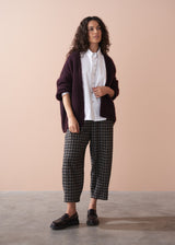 Woman wearing a dark purple cardigan, white shirt, plaid barrel leg trousers, and loafers against a beige background
