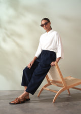 Woman in a white shirt and navy corduroy barrel leg trousers sitting on a wooden chair against a neutral background