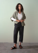 Woman wearing a green knitted shawl over a white shirt and dark jeans on a plain background