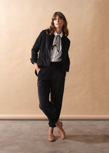 Woman wearing black corduroy trousers with a white shirt and patterned tie against a beige wall.