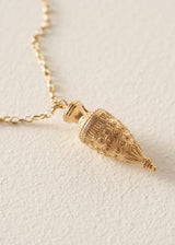 A gold-toned pendant necklace featuring an ornate, textured teardrop-shaped pendant on a delicate chain, set against a textured off-white background.