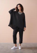 A woman wearing a dark grey oversized sweater over a striped top and black trousers with white trainers on a beige background