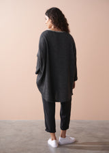 A woman wearing a dark grey oversized sweater over a striped top and black trousers with white trainers on a beige background