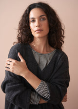 A woman wearing a dark grey oversized sweater over a striped top and black trousers with white trainers on a beige background