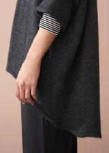 A woman wearing a dark grey oversized sweater over a striped top and black trousers with white trainers on a beige background