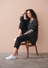 Woman sitting on a wooden chair wearing a dark grey jumper and black pants against a beige wall.