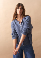 Woman wearing light blue wool blend cardigan with mother of pearl buttons and wide leg jeans against a beige background
