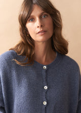 Close up of model wearing light blue wool blend cardigan with mother of pearl buttons and wide leg jeans against a plain background
