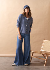 A model in a light blue wool blend cardigan, mother of pearl buttons, wide-leg jeans, and a denim cap stands next to a chair against a plain background.