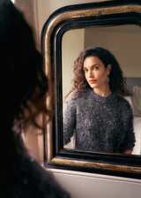 Woman wearing a dark grey textured sweater with sequins and statement earrings, looking into an antique mirror