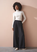 Woman wearing a white turtleneck and black wide-leg pants against a beige wall.