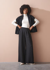 Person wearing a white turtleneck, black vest, and wide-leg pants against a beige wall.