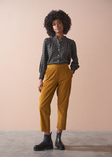 Woman wearing a black checked shirt and mustard corduroy trousers against a beige background
