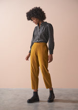Woman wearing a black checked shirt and mustard corduroy trousers against a beige background