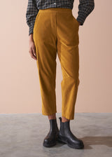 Woman wearing a black checked shirt and mustard corduroy trousers against a beige background