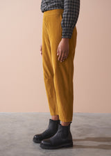 Woman wearing a black checked shirt and mustard corduroy trousers against a beige background