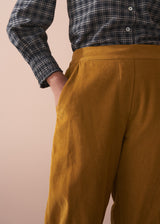 Woman wearing a black checked shirt and mustard corduroy trousers against a beige background