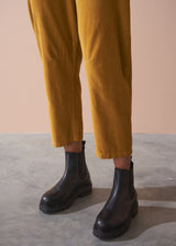 Woman wearing a black checked shirt and mustard corduroy trousers against a beige background