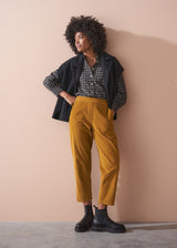 Woman wearing a black checked shirt and mustard corduroy trousers against a beige background