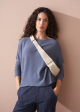 Woman wearing a grey sweater and dark pants with a beige crossbody bag against a beige wall.