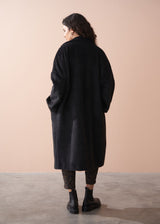 Person wearing a black coat standing against a beige wall.